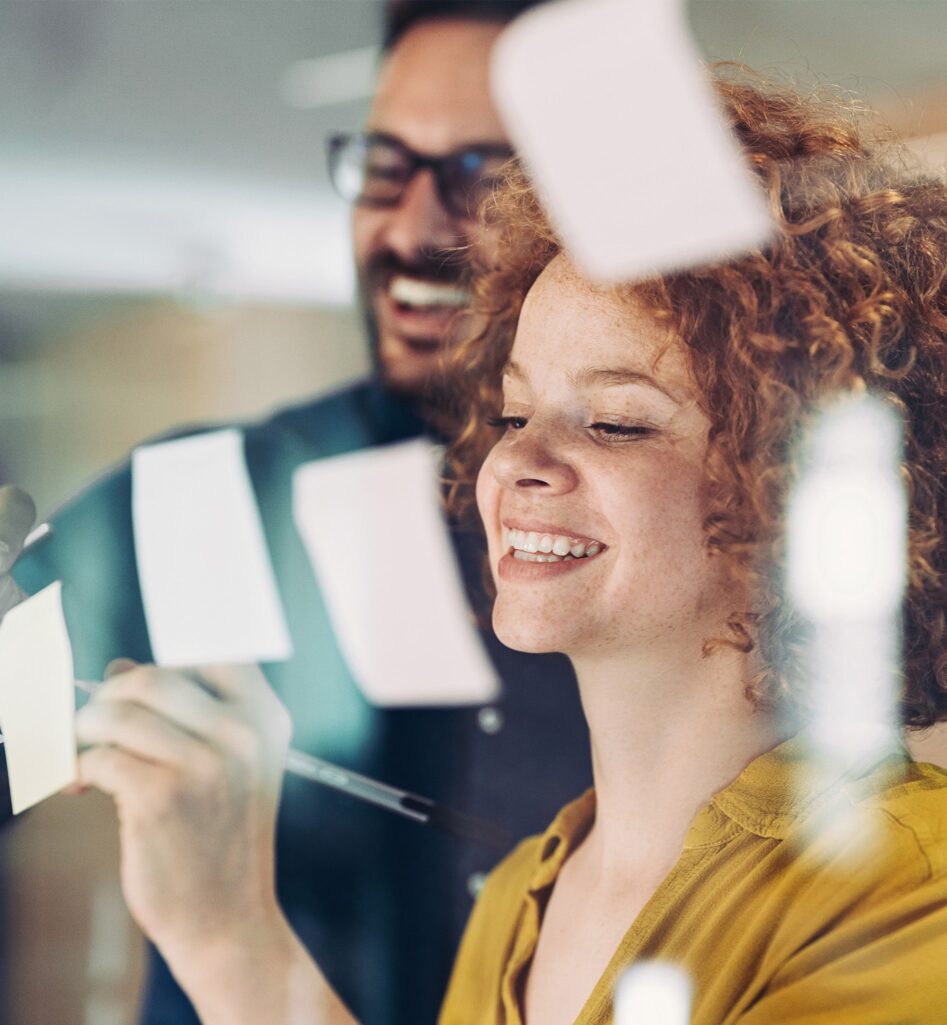 Een vrouw en een man schrijven op post-it briefjes die voor een raam hangen.