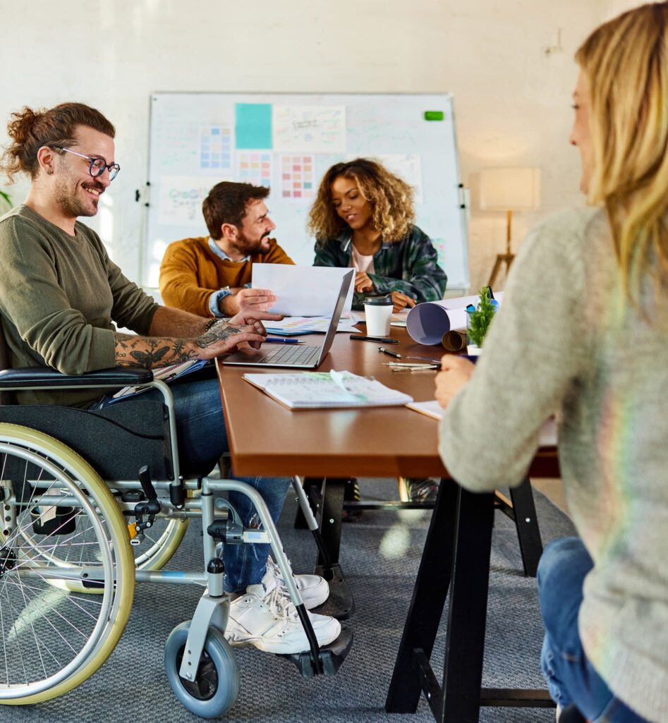 Een man in een rolstoel werkt aan zijn computer met drie andere mensen