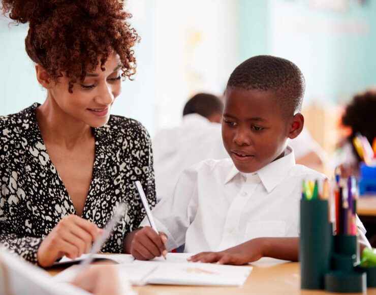 Woman Elementary School Teacher Giving Male Pupil Wearing Uniform One To One Support In Classroom pour illustrer US Lusitanos, club de football, association soutenue par le groupe Blue Soft dans le cadre du Projet Indigo 2026